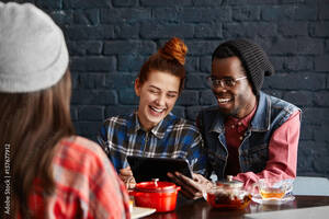 black funny videos - Handsome African student wearing stylish black hat holding touch pad,  showing funny videos on internet to his beautiful redhead girlfriend who is  looking at screen of digital tablet and laughing Stock Photo |